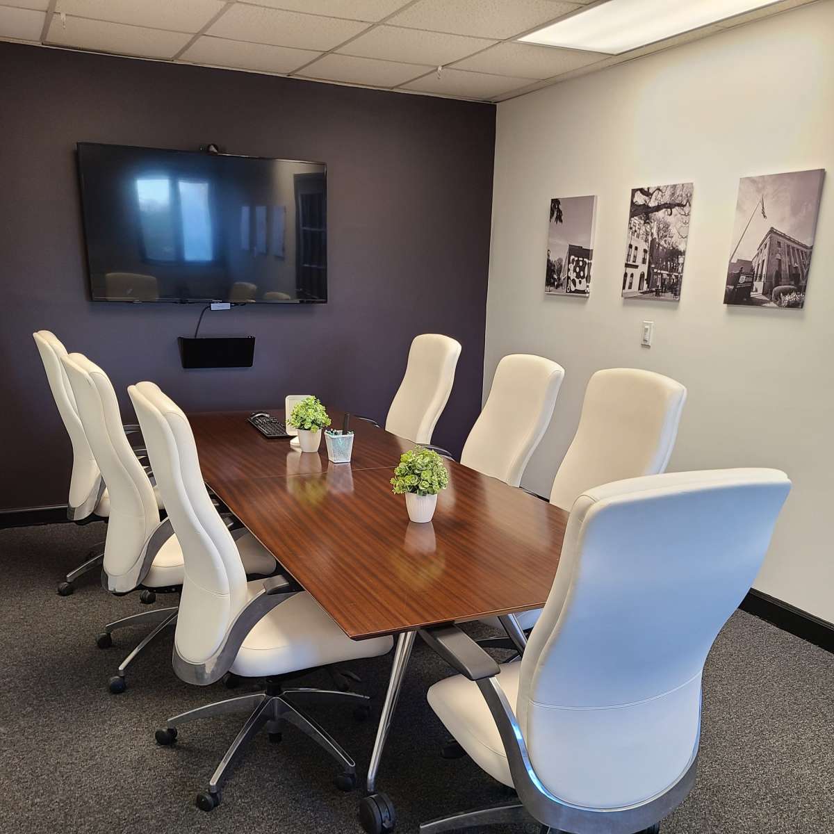 A modern conference room features a large wooden table surrounded by eight white ergonomic chairs and a wall-mounted television.