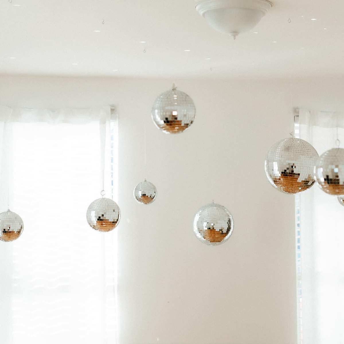 Several disco balls hanging from the ceiling in a room with white walls and sheer curtains.
