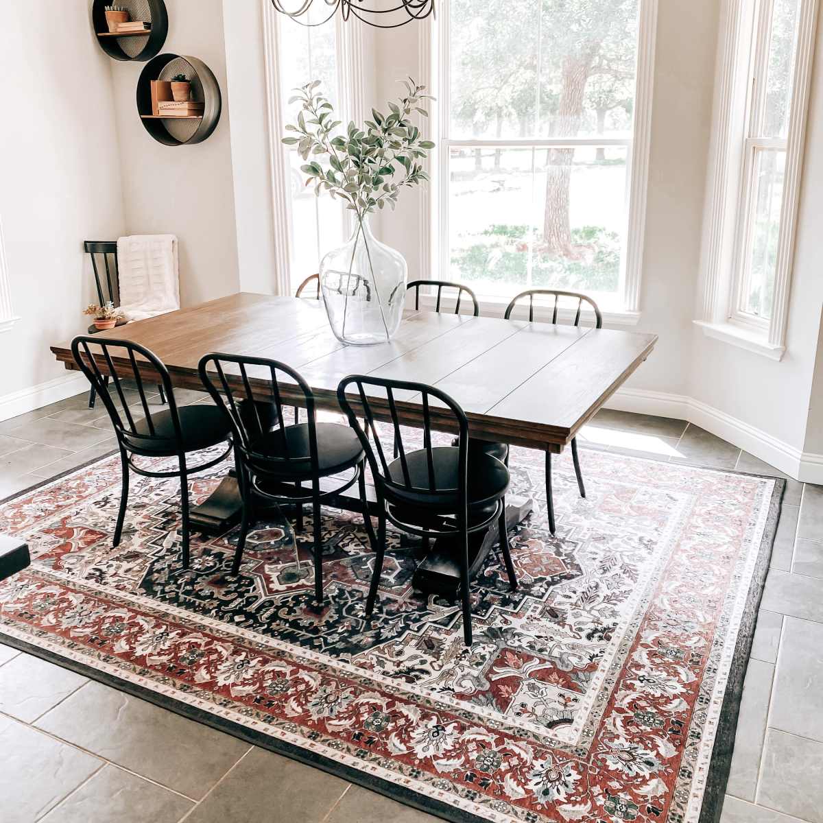A dining room features a wooden table surrounded by black chairs on a patterned rug, with large windows allowing natural light to fill the space.