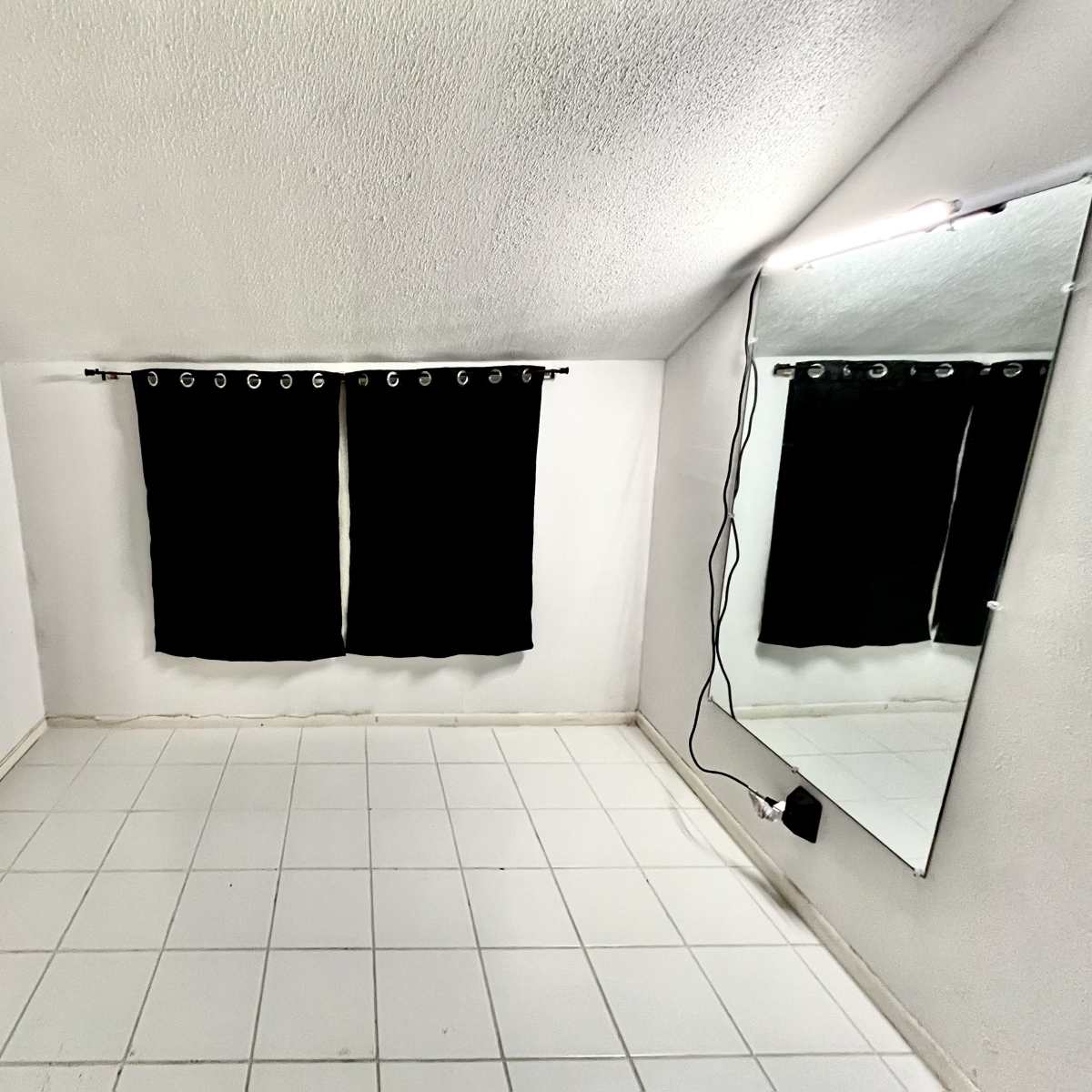 A sparsely furnished white room with two black curtains on a window and a large mirror mounted on the wall.