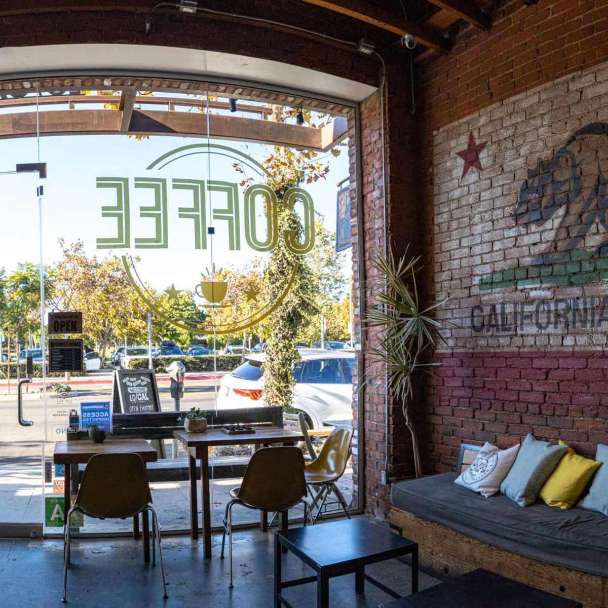 A coffee shop interior featuring a large mural of a bear and the words "California Republic" on a brick wall, with tables and chairs arranged near a glass entrance.