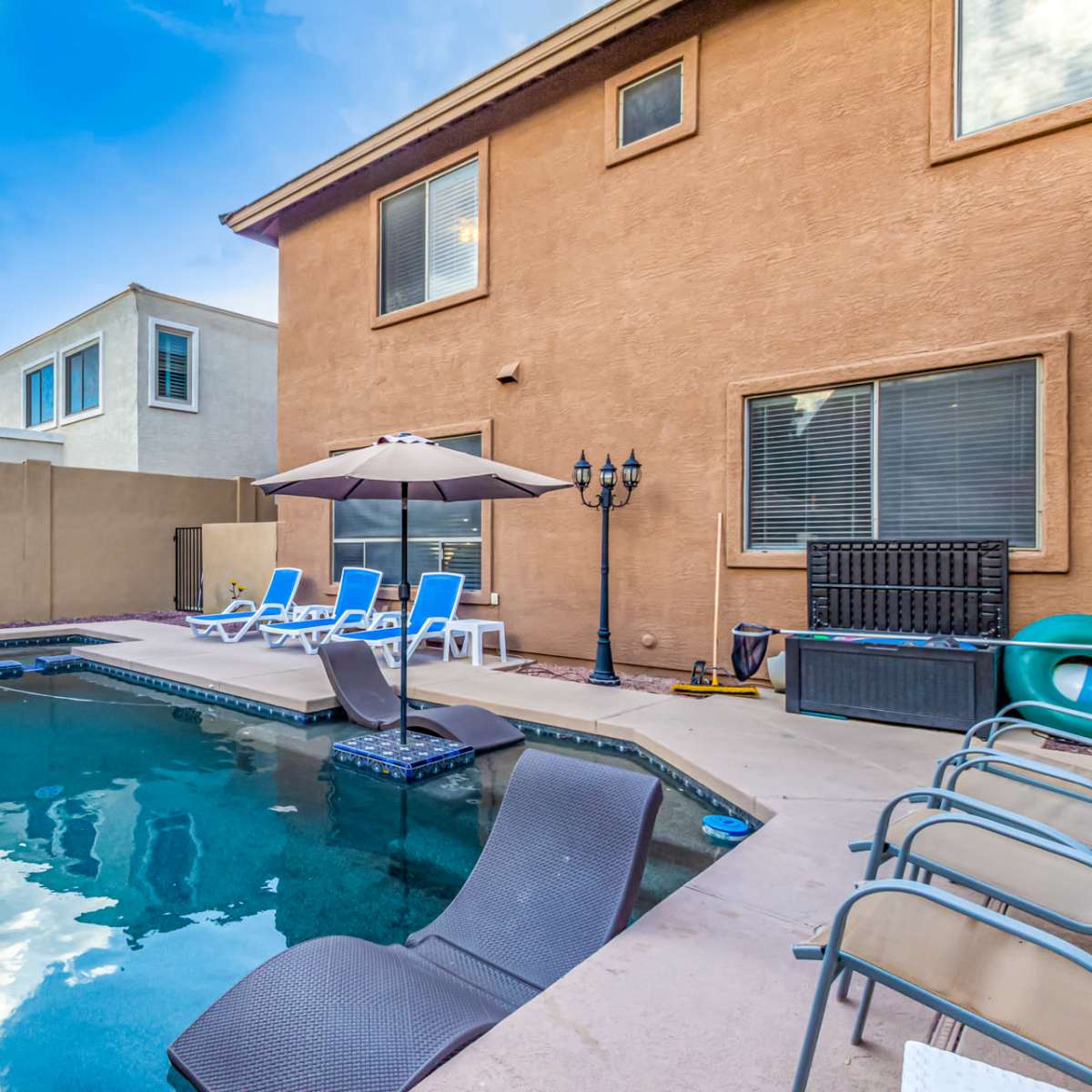 A swimming pool area features lounge chairs and an umbrella, surrounded by a beige wall and a two-story building.