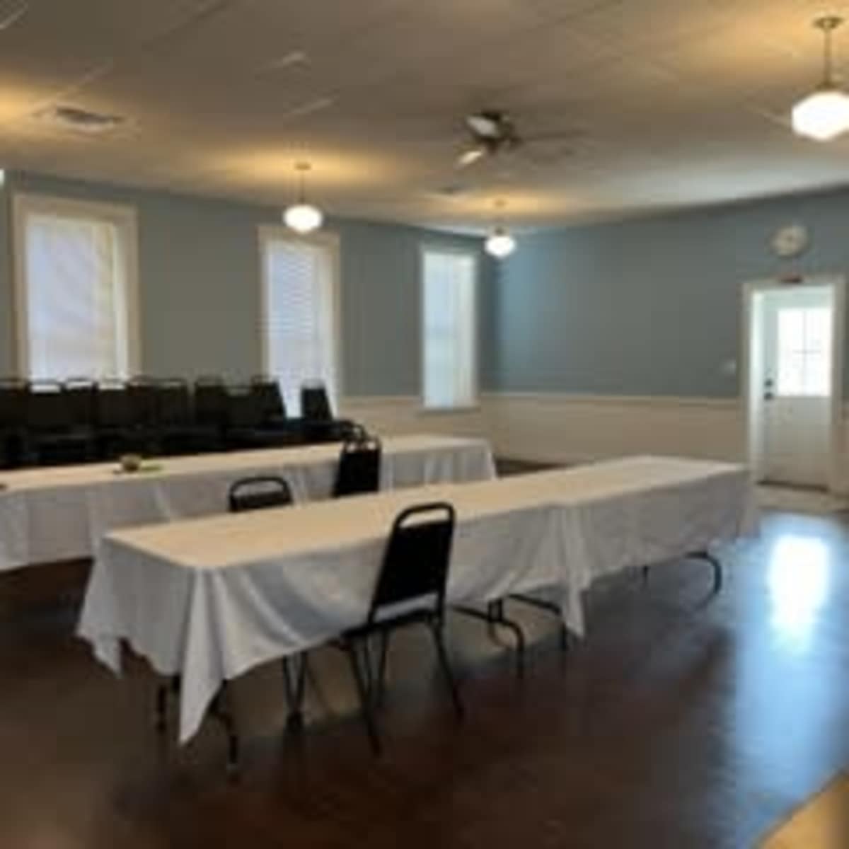 A meeting room with two long tables covered in white tablecloths, black chairs stacked against the wall, and a door leading outside.