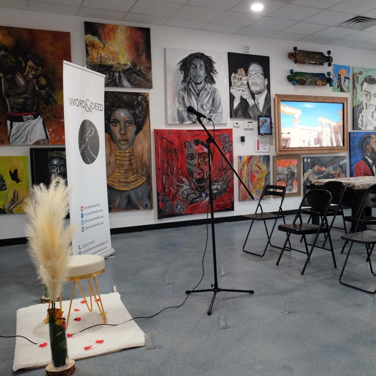 A room set up for an event, featuring empty chairs, a microphone stand, and various colorful artworks on the walls.