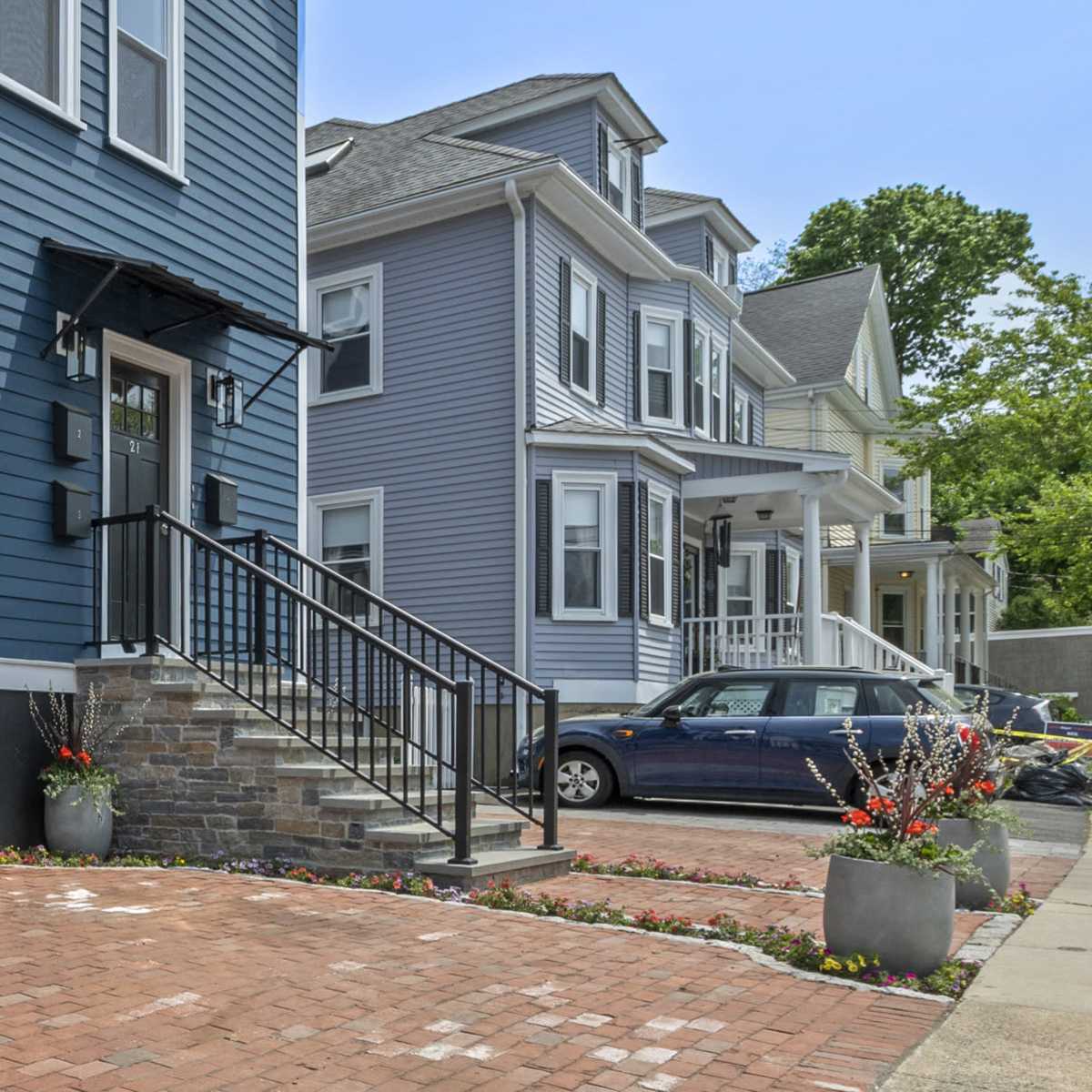 A row of residential buildings with blue and gray siding, a brick walkway, and planters in front, set against a clear sky.