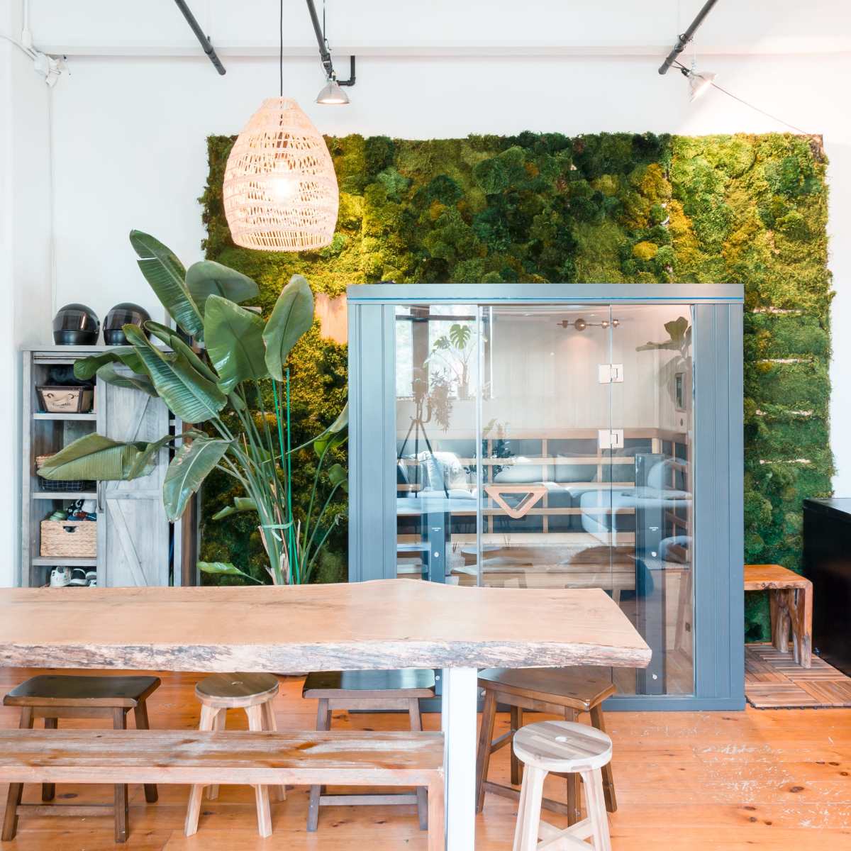 A spacious, modern interior features a wooden dining table, a glass-enclosed kitchenette, and a moss wall backdrop with tropical plants.