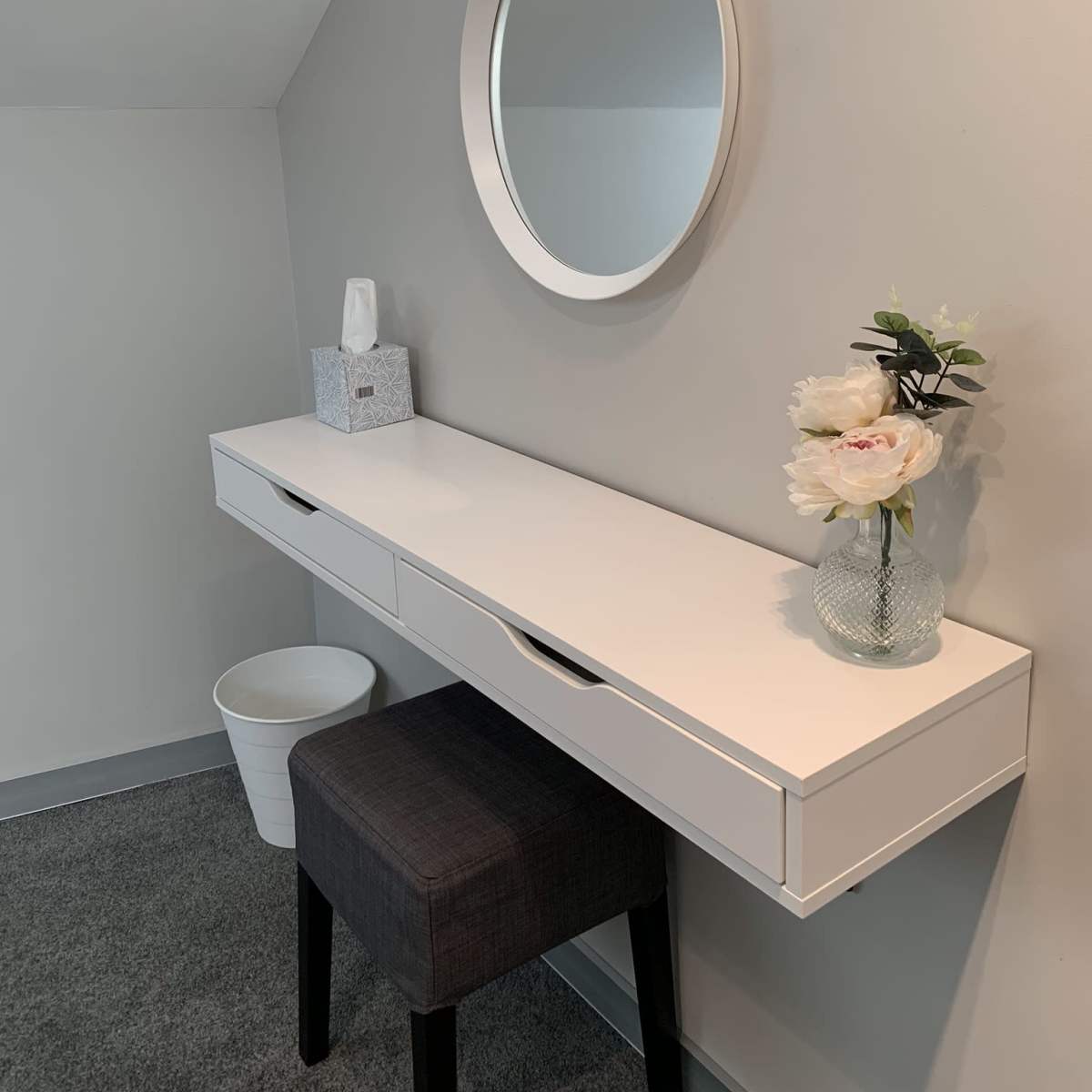 A minimalist vanity table with a round mirror, a vase of flowers, and a tissue box against a light gray wall.