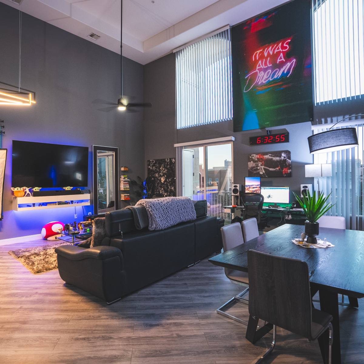 A modern living space with a black sofa, a dining table, and a wall-mounted television, illuminated by ambient lighting and large windows.