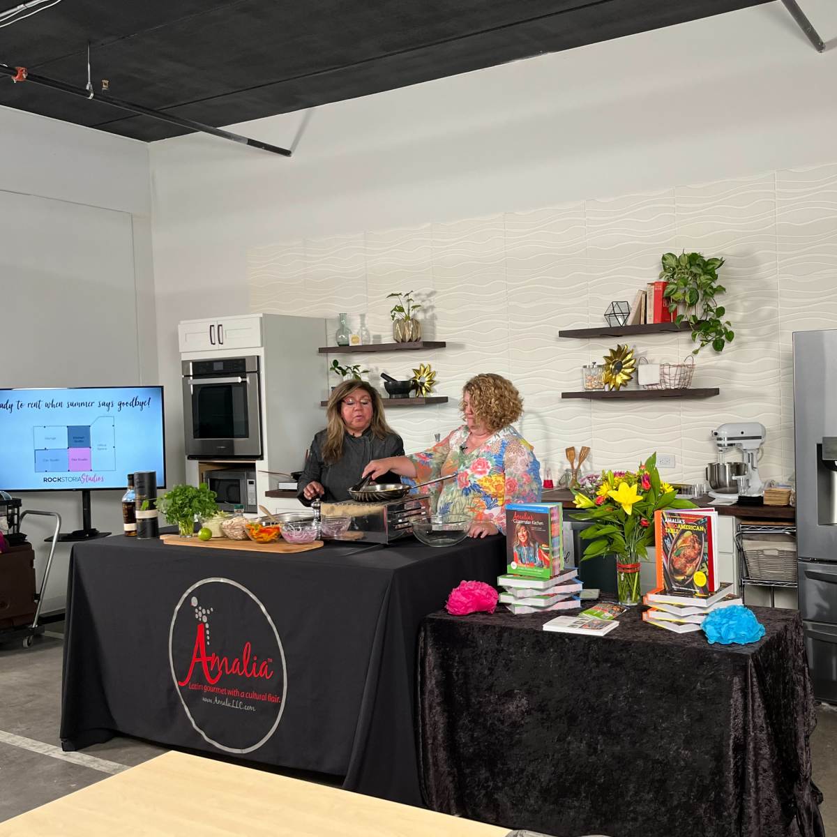 Two women are preparing food at a kitchen counter decorated with plants and cookbooks, while a large sign welcomes visitors to the cooking space.