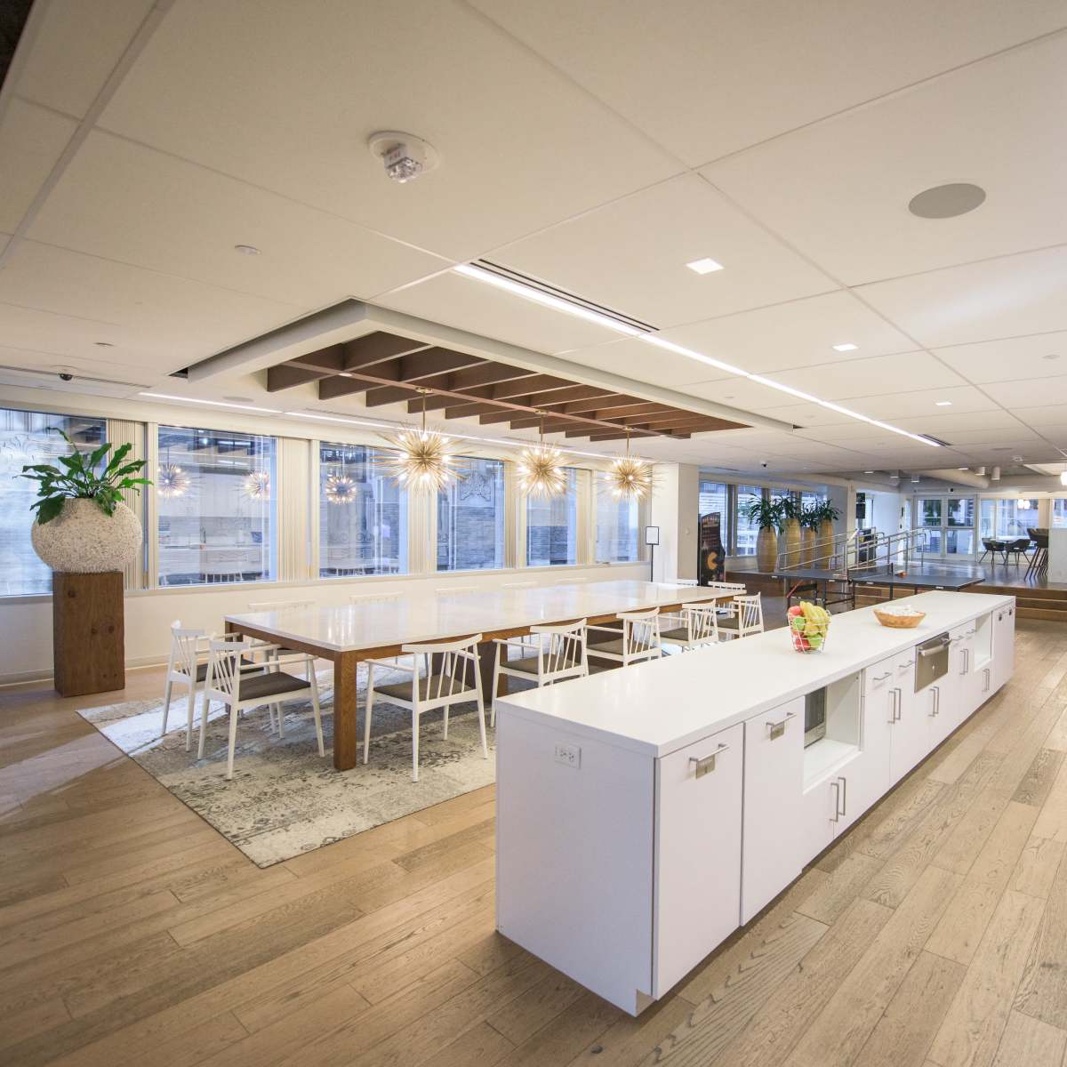 A modern office kitchen and dining area with a long white table, surrounding chairs, and large windows letting in natural light.