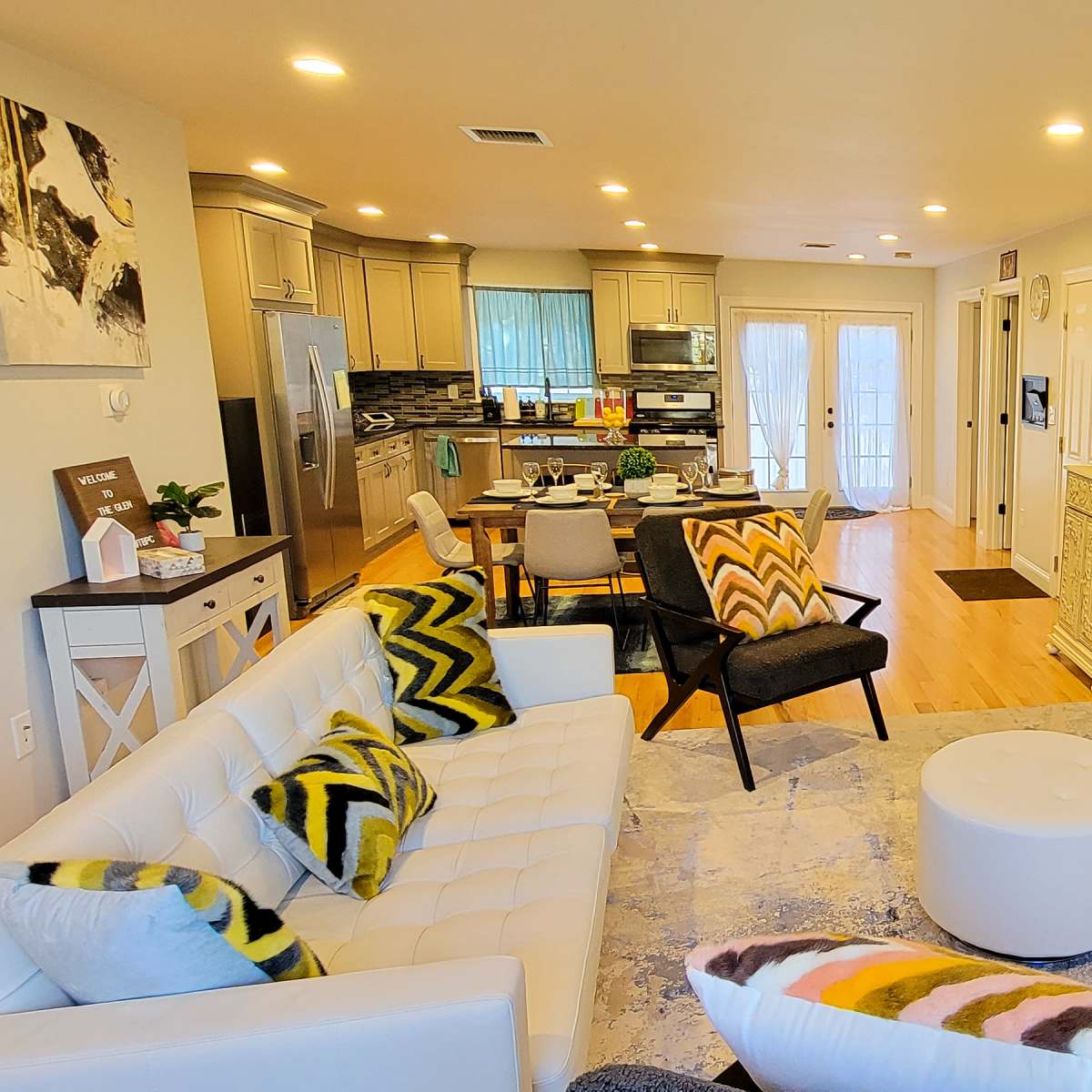 A modern living room with a white sectional sofa, decorative cushions, a wall-mounted television, and an open layout leading to a kitchen area.