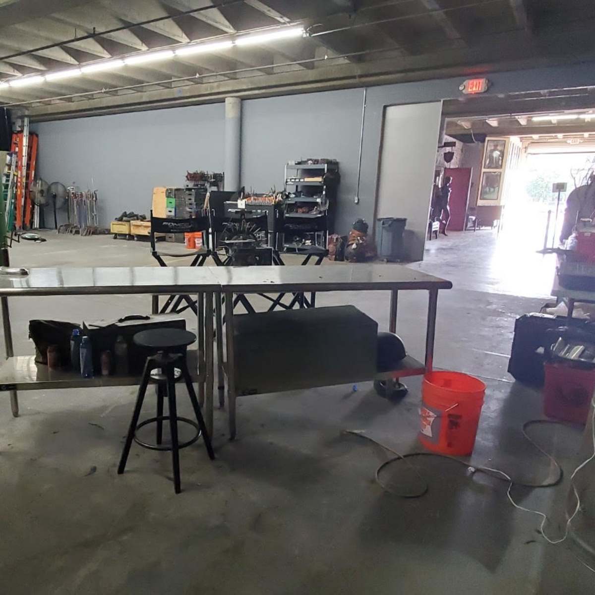 An industrial workspace featuring a metal table, a stool, a large orange bucket, and various tools and equipment along the walls.
