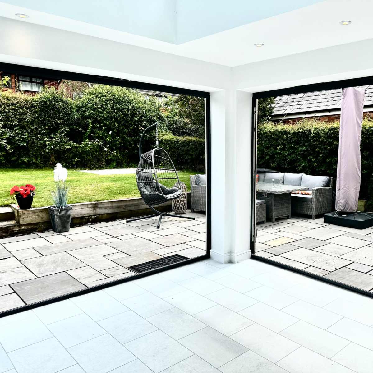 A modern interior space with large open doors that lead to a stone-paved patio and a well-maintained garden.