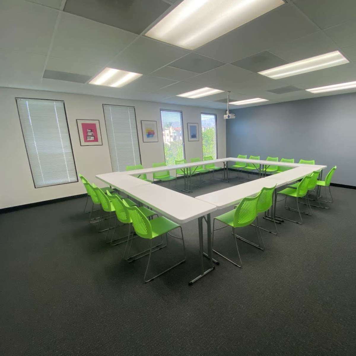 A modern meeting room features a square arrangement of white tables surrounded by green chairs, with large windows letting in natural light.