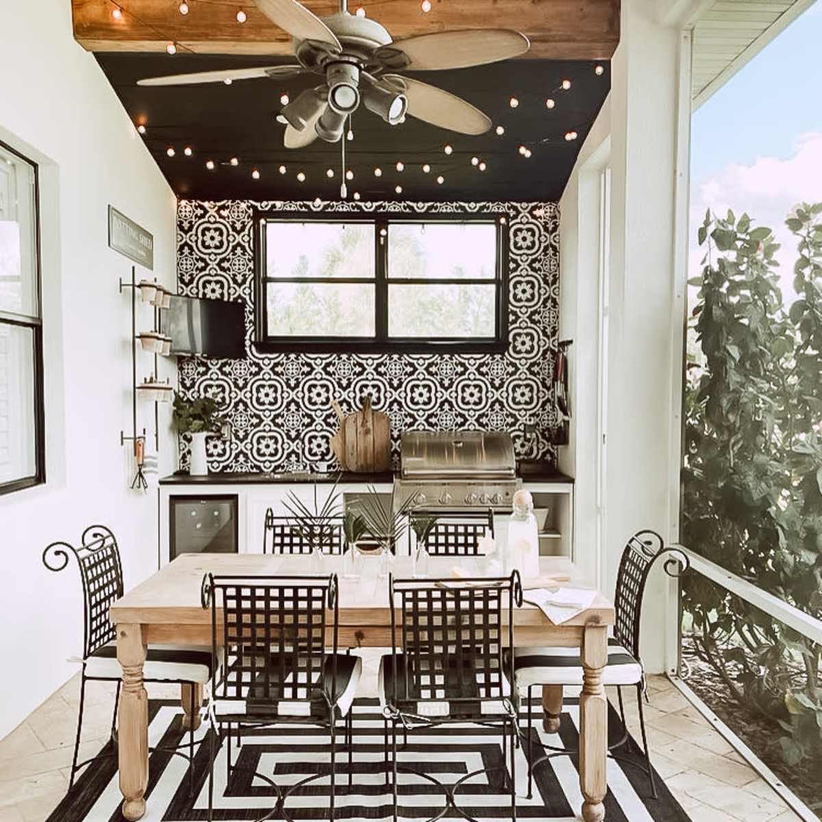 An outdoor dining area with a wooden table, black metal chairs, and an outdoor grill, surrounded by patterned tiles and string lights.