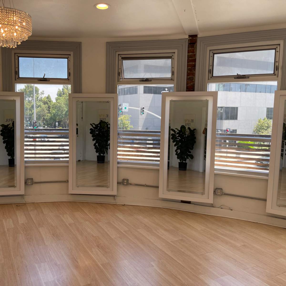 A bright room with large mirrors and a chandelier, featuring potted plants in each mirror's reflection.