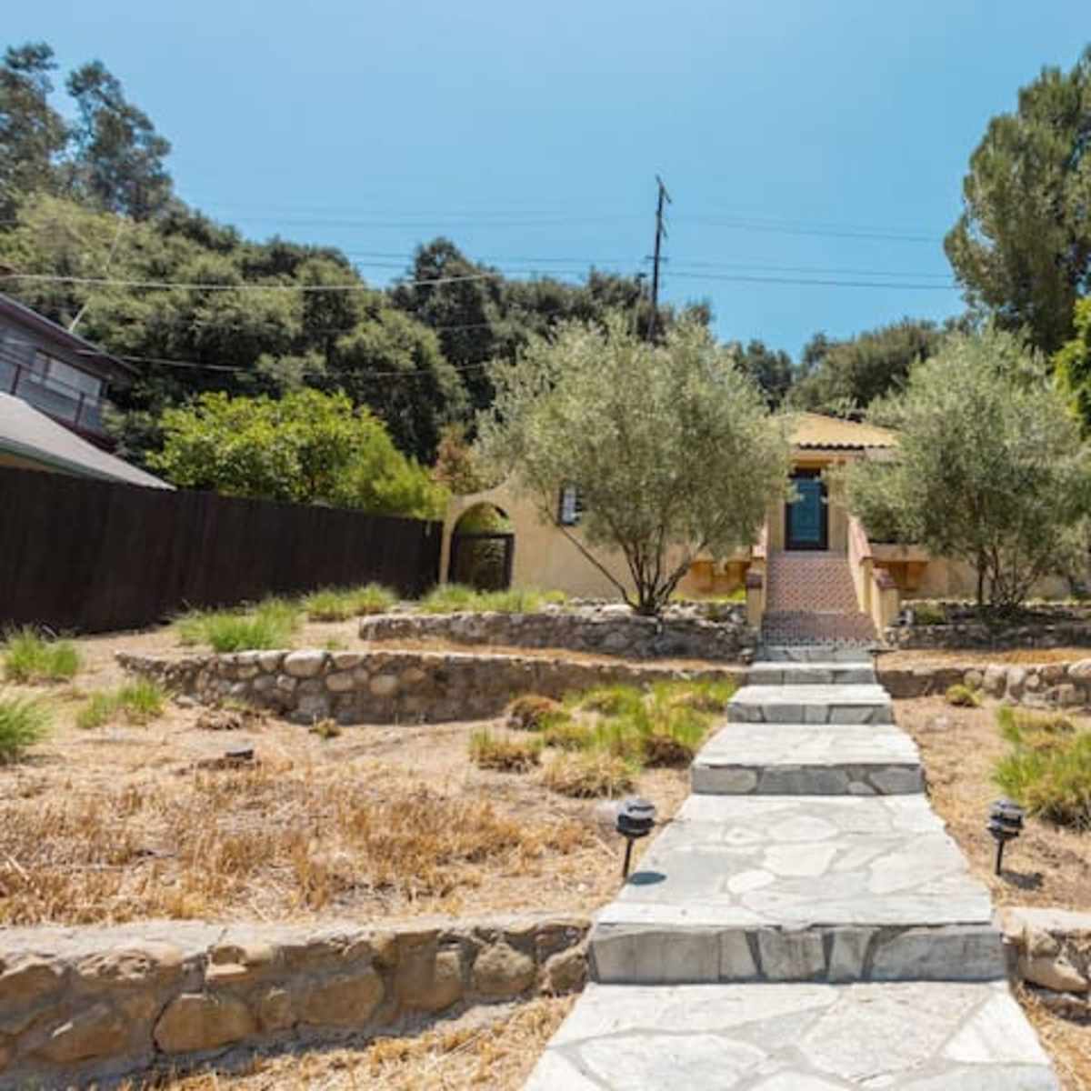 A stone pathway leads to a yellow house surrounded by olive trees and dry landscaping.