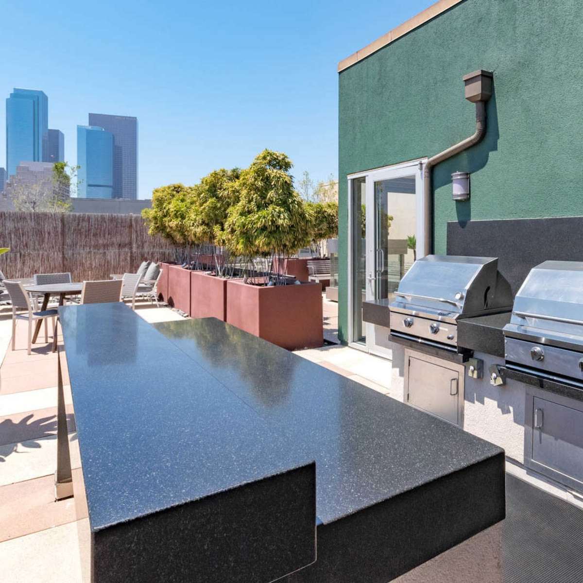 An outdoor kitchen area with two stainless steel grills and a long black countertop, set against a backdrop of downtown Los Angeles skyscrapers.