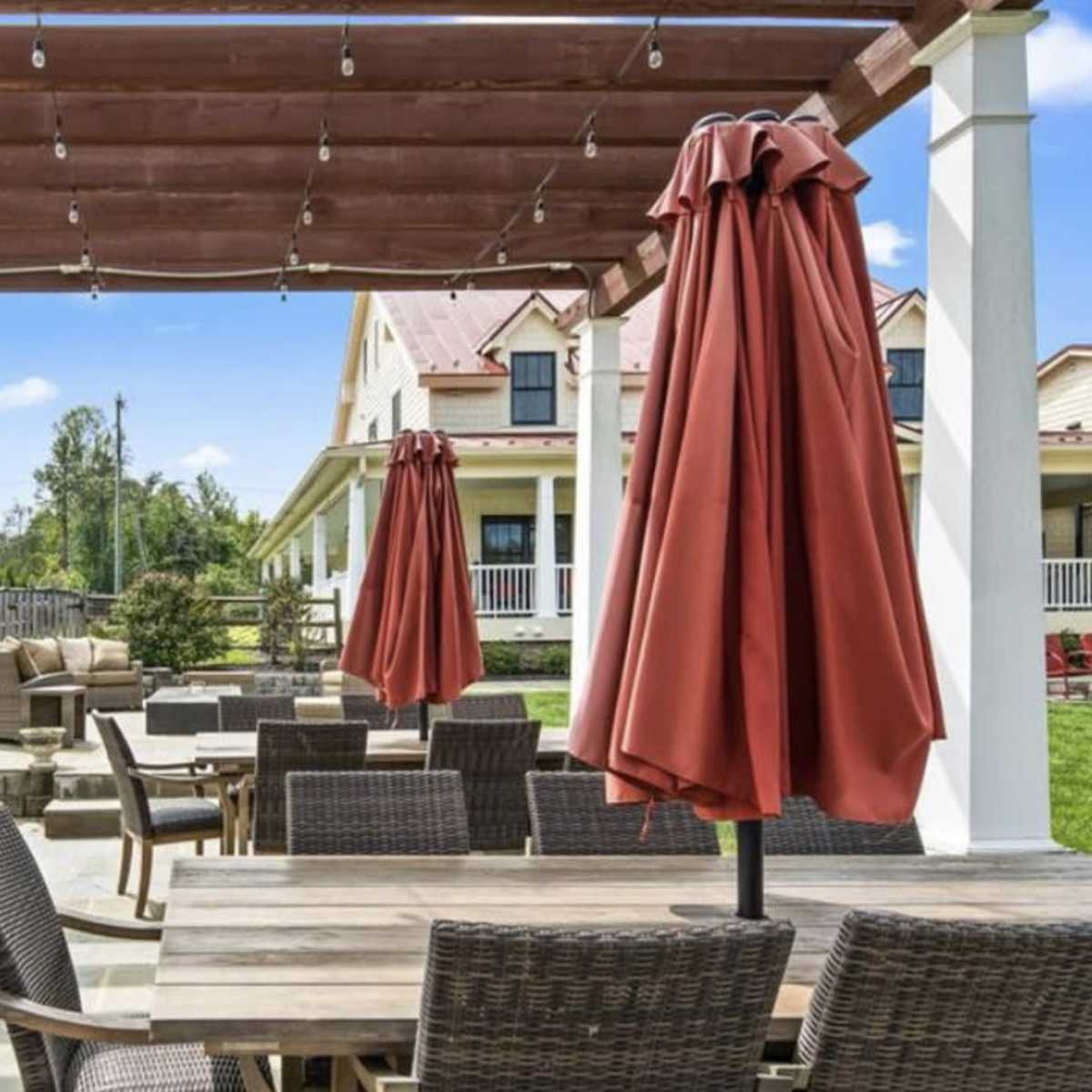 An outdoor patio with a wooden pergola, seating arrangements, and a grill, surrounded by well-maintained greenery.