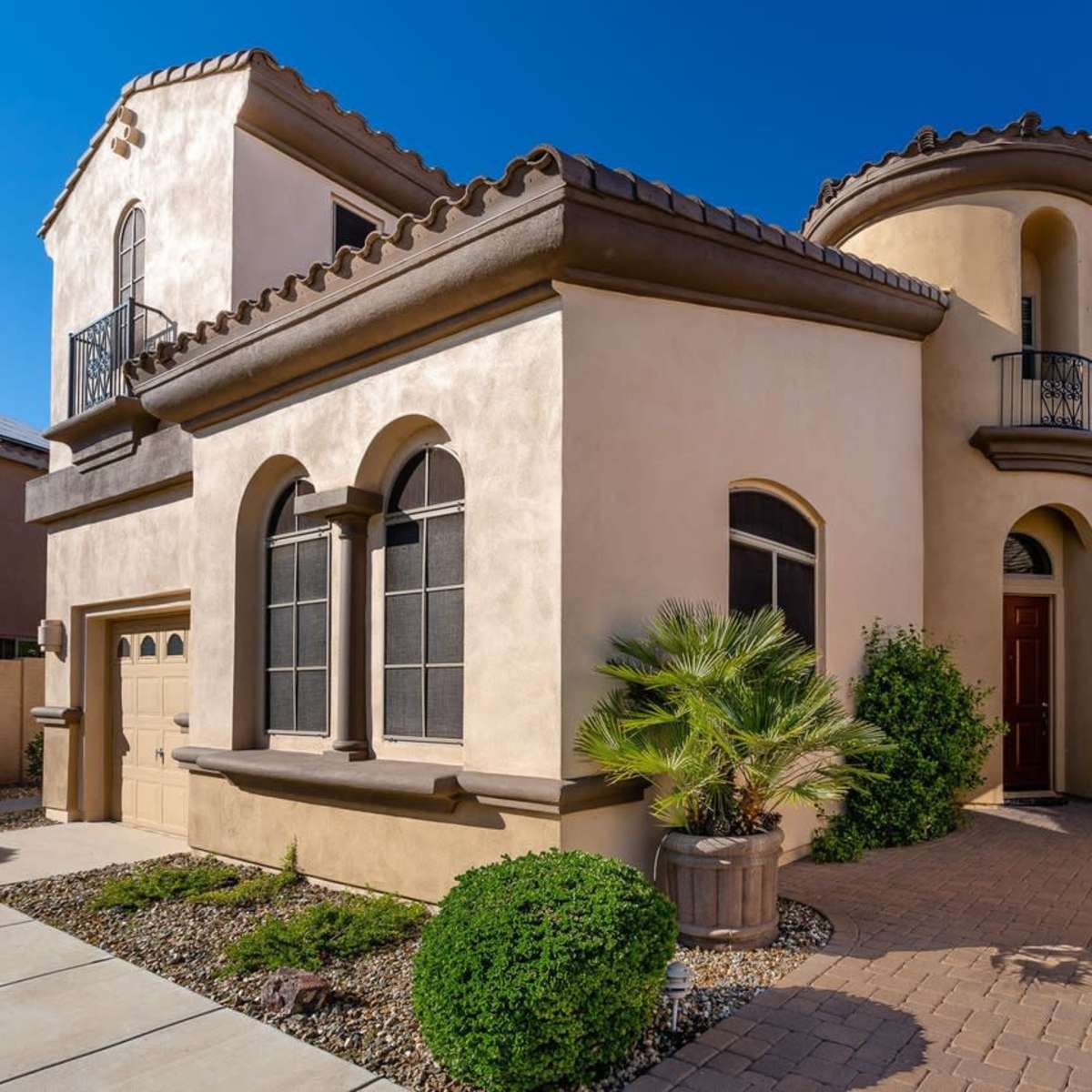 A two-story house with a rounded turret, arched windows, and palm trees in the front yard, surrounded by paved pathways.