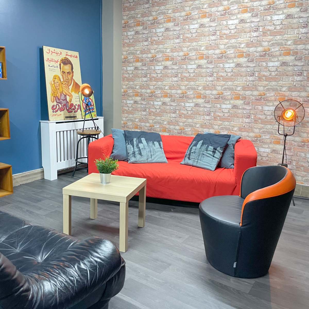 A cozy living room with a red couch, a black leather chair, a small beige coffee table, and a decorative bookshelf against a blue wall with a brick accent.
