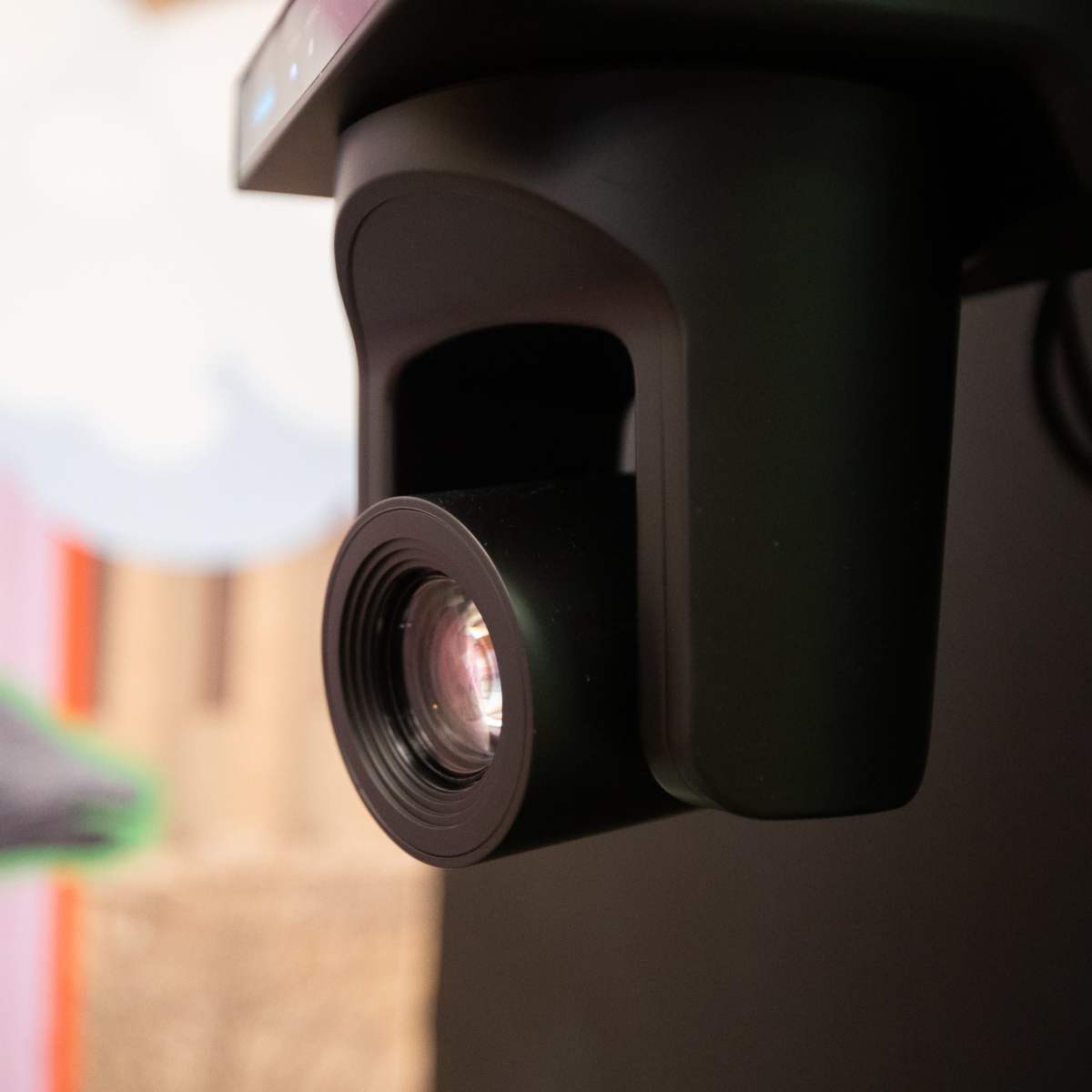 A close-up of a black camera lens mounted on a device, with a colorful background featuring stylized clouds and a hand.