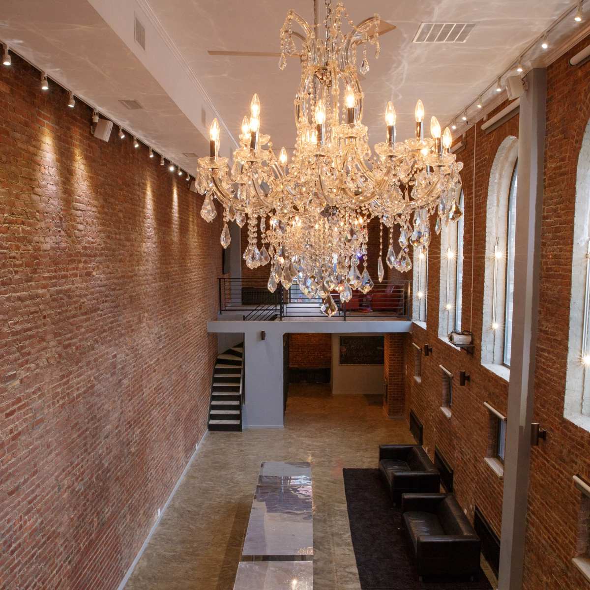A spacious interior features a large chandelier suspended from the ceiling, with exposed brick walls and a polished floor, complemented by black sofas along the sides.