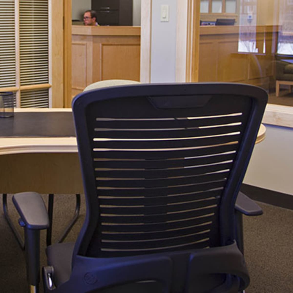 An empty office with a curved desk, a black ergonomic chair, a telephone, and a background area with a person visible through a glass partition.