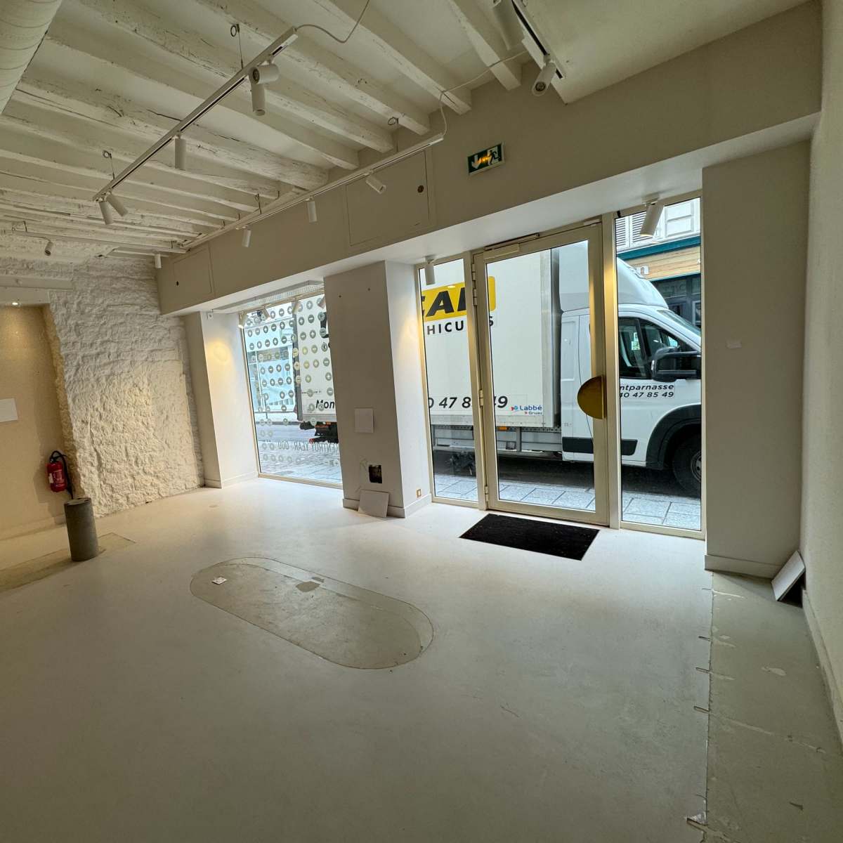 An empty commercial space with a stone wall, white ceiling beams, and large glass doors facing a street, while a moving truck is parked outside.
