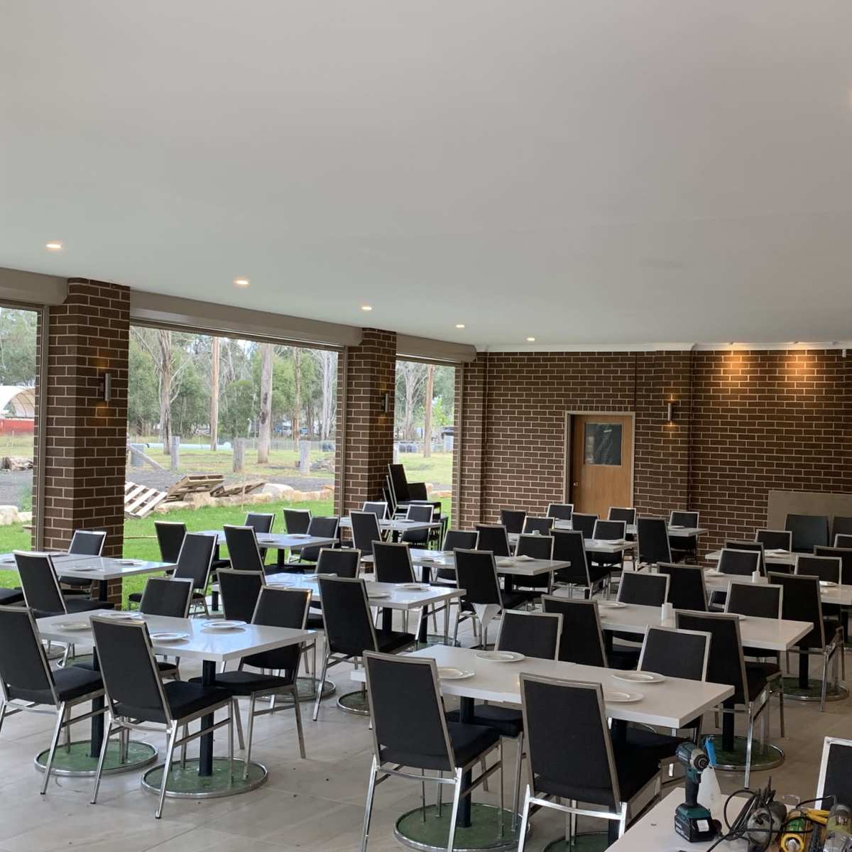 A spacious dining area with multiple tables and chairs arranged systematically, and large windows providing a view of the outside greenery.