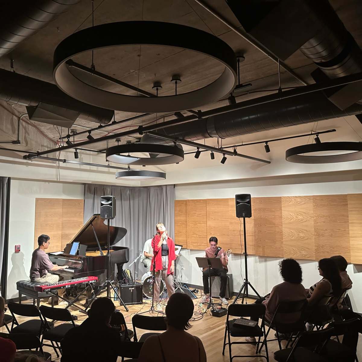 A small music performance features a singer and three musicians playing a piano, keyboard, and drums in a well-lit room with chairs arranged for the audience.
