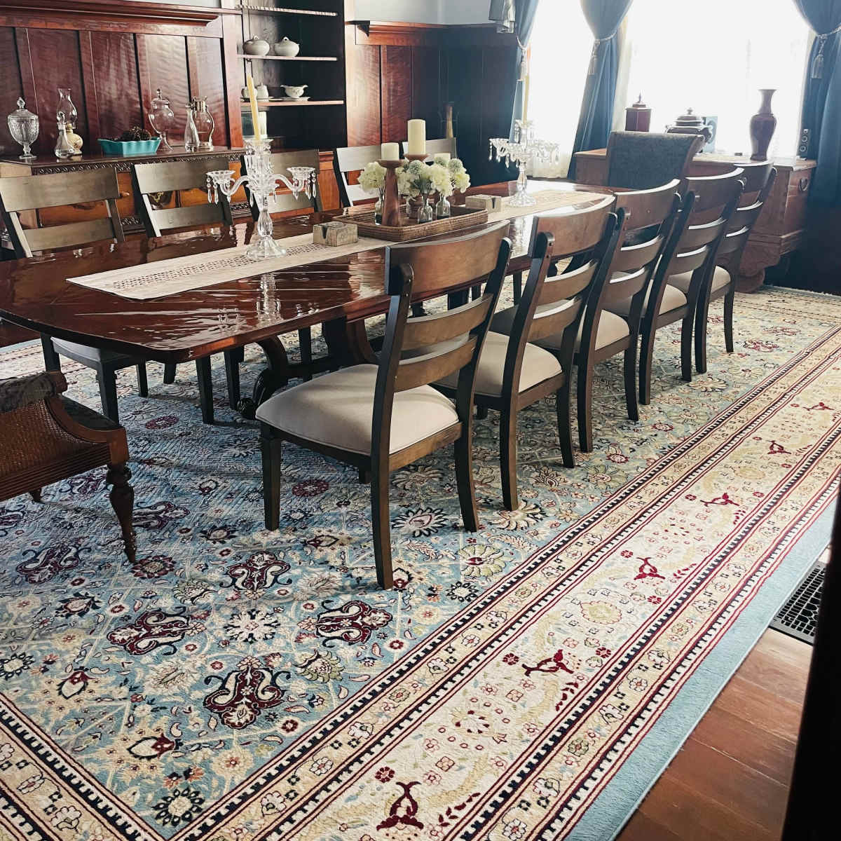 A large dining room features a polished wooden table surrounded by ten chairs, a patterned rug, and elegant decor along the walls.