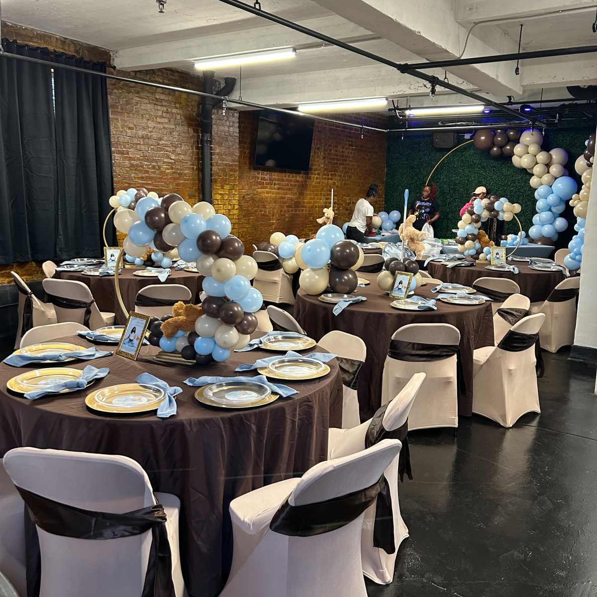 A decorated event space featuring several round tables covered with brown cloths, set with plates and balloons in shades of blue and brown.