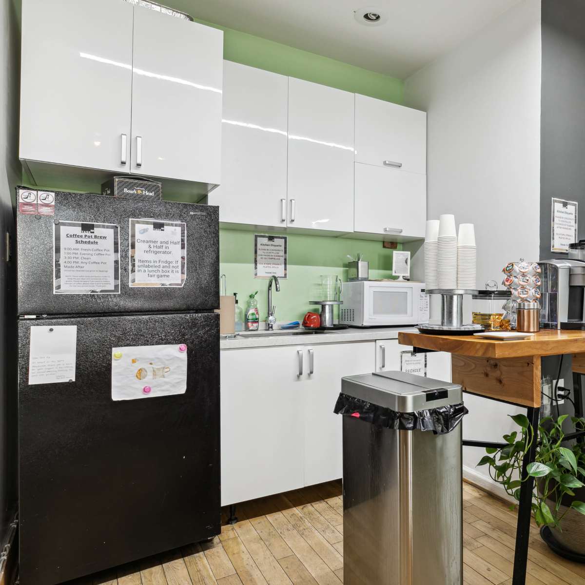 A small kitchen area with a white refrigerator, a microwave, and a wooden table, accompanied by a trash bin and potted plant.