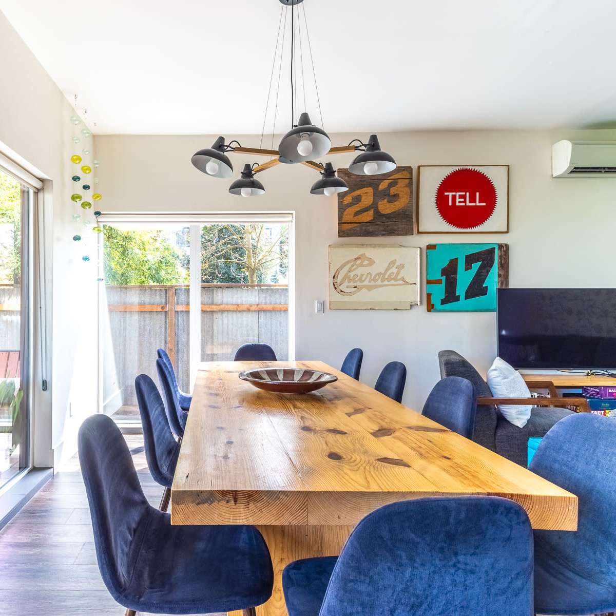 A large wooden dining table surrounded by blue chairs is illuminated by pendant lights in a bright dining area with artwork on the walls.