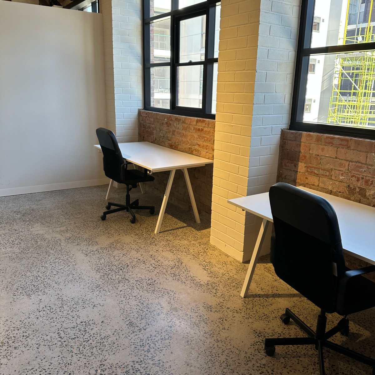 A light-filled office space featuring two desks with black chairs situated near large windows, against a backdrop of exposed brick walls.