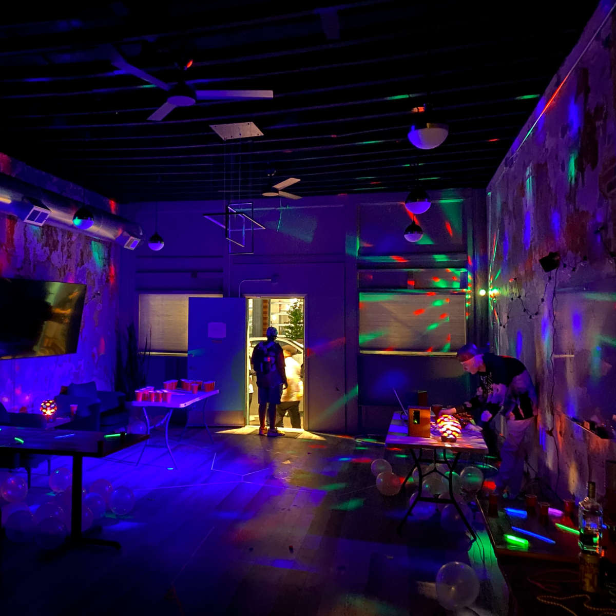 A dimly lit room features colorful lights reflecting off the walls, with party decorations and balloons scattered across the floor.