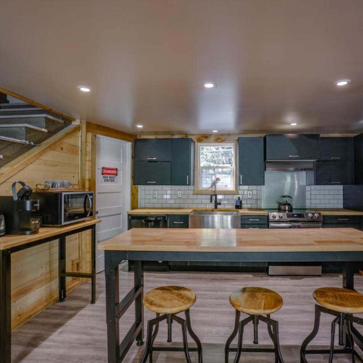 A modern kitchen features wooden cabinetry, stainless steel appliances, a central island with bar stools, and a staircase in the background.