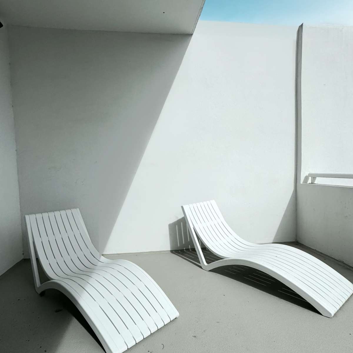 Two white lounge chairs are positioned on a smooth concrete surface beneath a minimalist white wall, with a clear blue sky visible in the background.