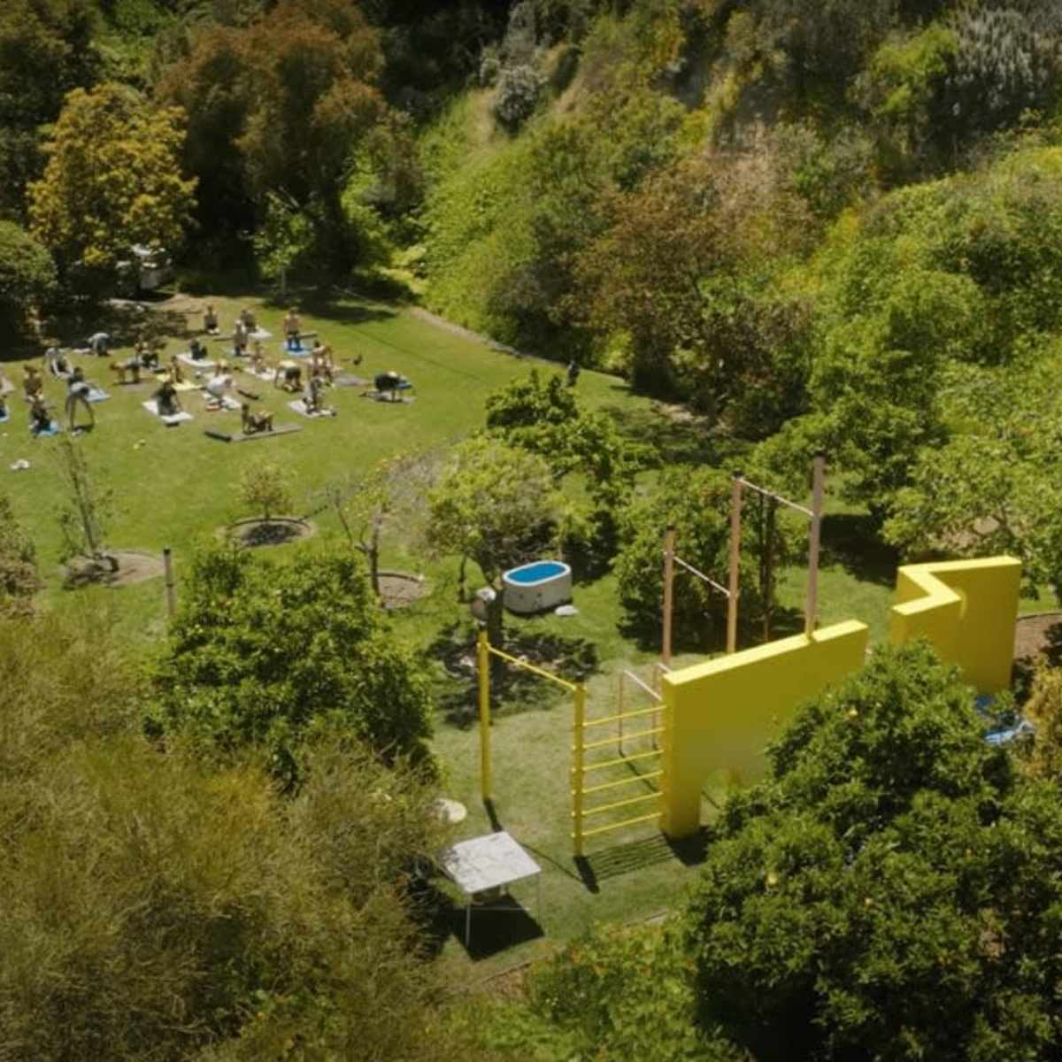 A large green outdoor space with various exercise equipment and people scattered around engaging in activities, surrounded by trees and hills.