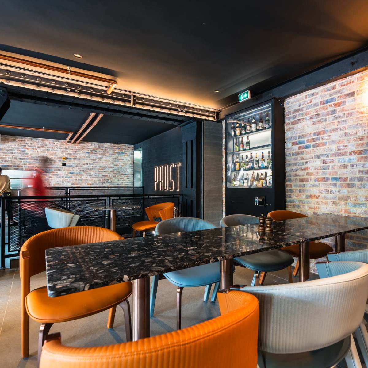 A modern dining area with a long black table surrounded by colorful chairs, set against a brick wall and industrial-style decor.