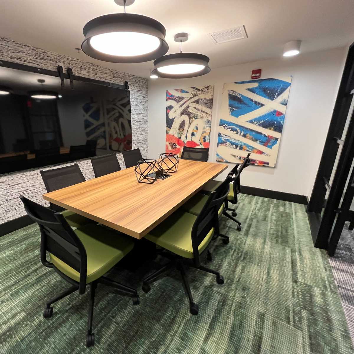 A modern conference room features a long wooden table surrounded by black chairs with green accents, illuminated by pendant lights, and includes a large mirror and abstract artworks on the walls.