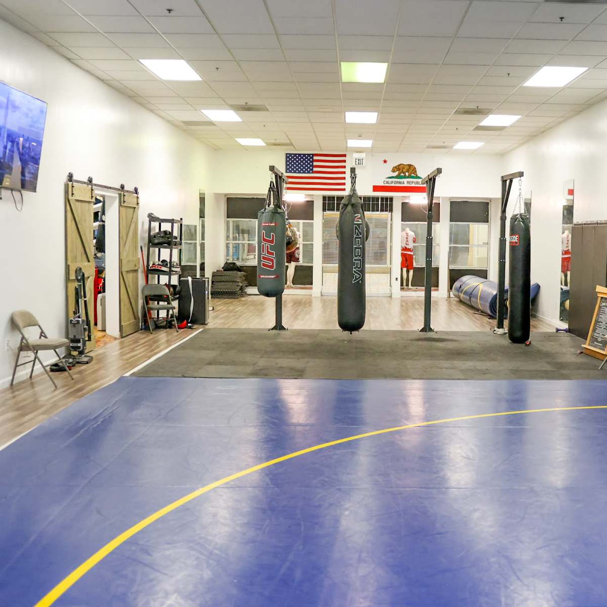 An empty fitness facility featuring punching bags, a wrestling mat, and workout equipment along the walls.