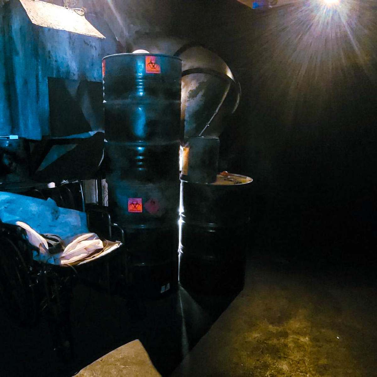 A dimly lit industrial space featuring several black barrels and a metal workbench against a dark wall.