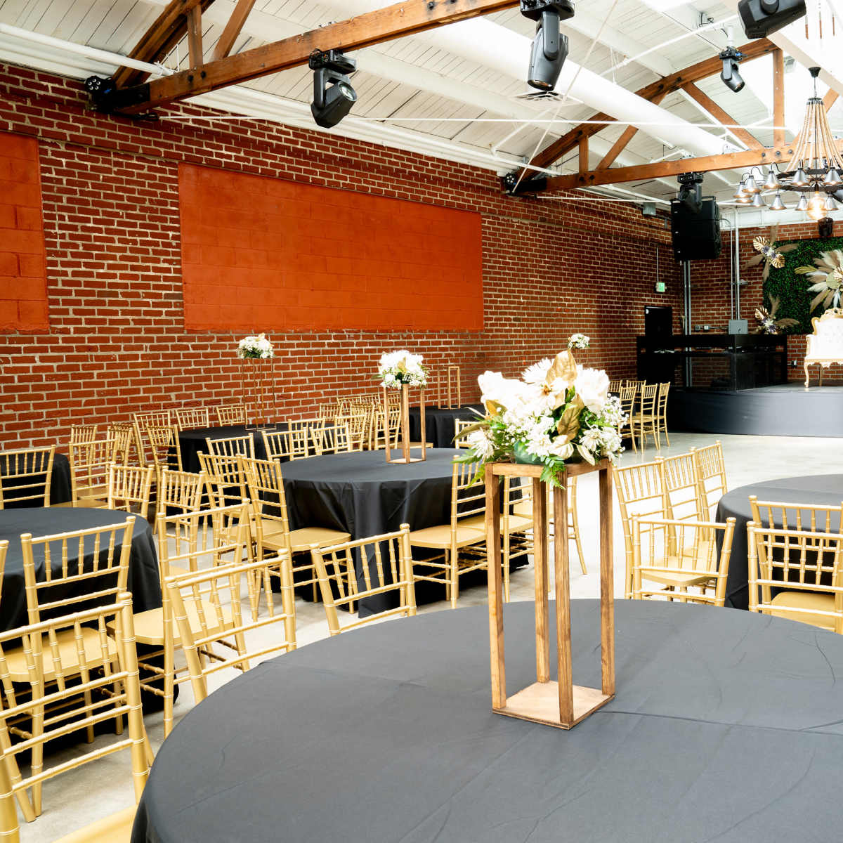 A spacious event venue with round tables covered in black tablecloths, decorated with floral centerpieces, and a stage at the front featuring elegant chairs and wall decor.