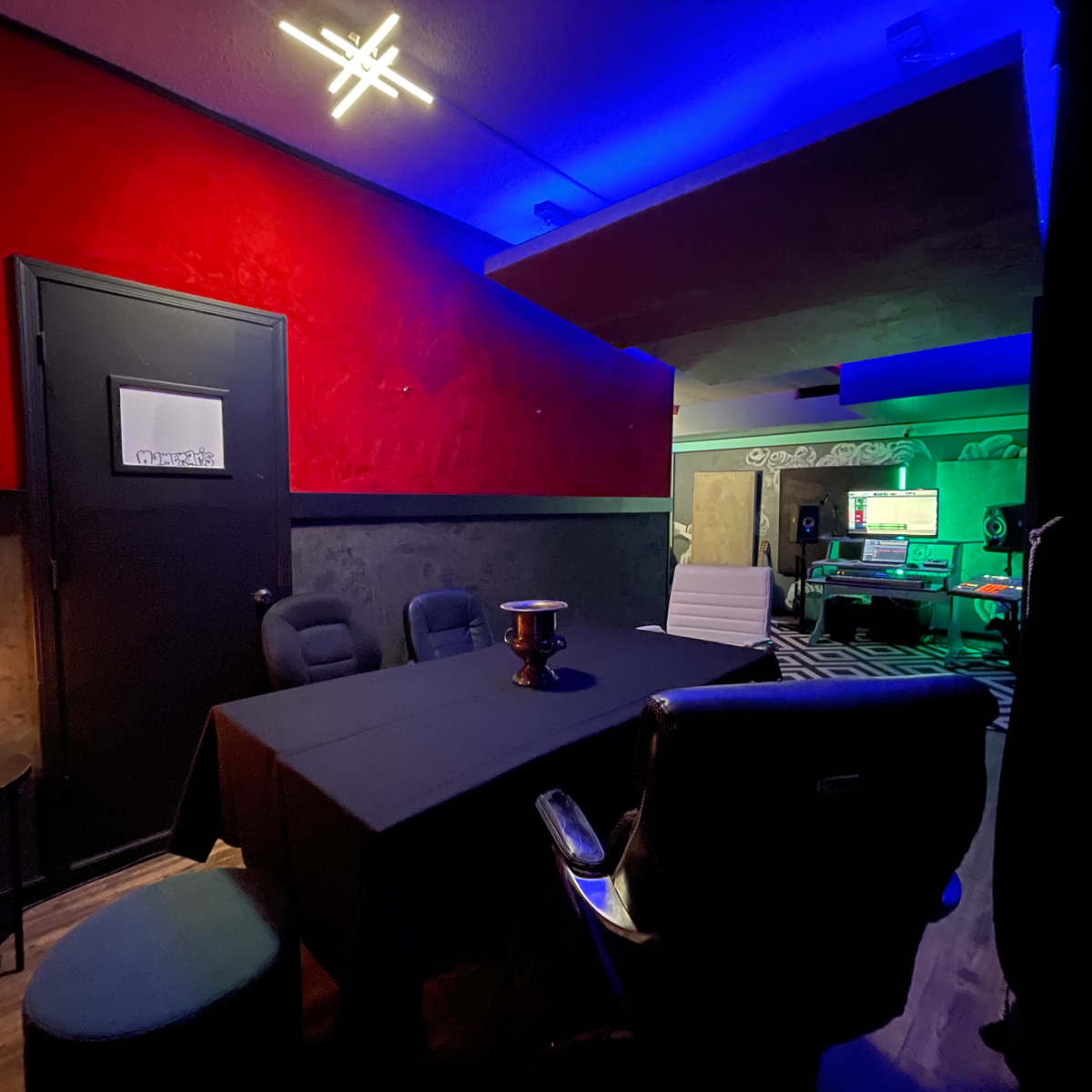 A dimly lit room features a black table surrounded by chairs, with a red accent wall and blue lighting in the background.