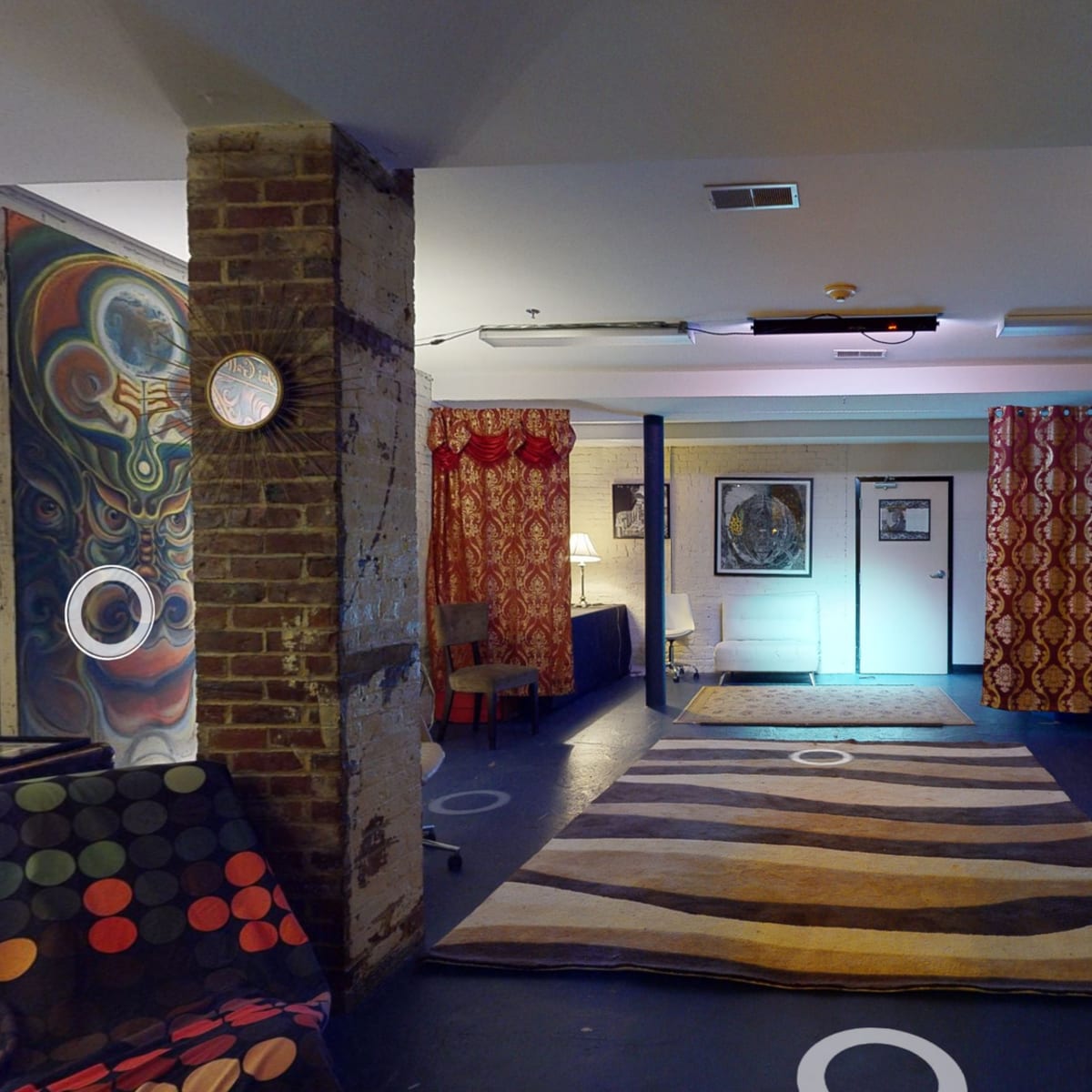 A spacious, lit room with patterned curtains, a striped rug, and artwork on the walls, featuring two brick columns.