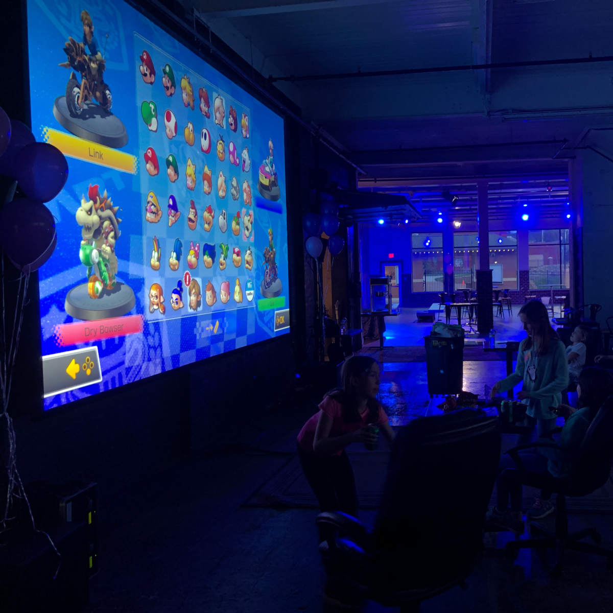 The scene shows a gaming event in a spacious room with a large screen displaying characters from a racing game, while children sit in gaming chairs in front of it.
