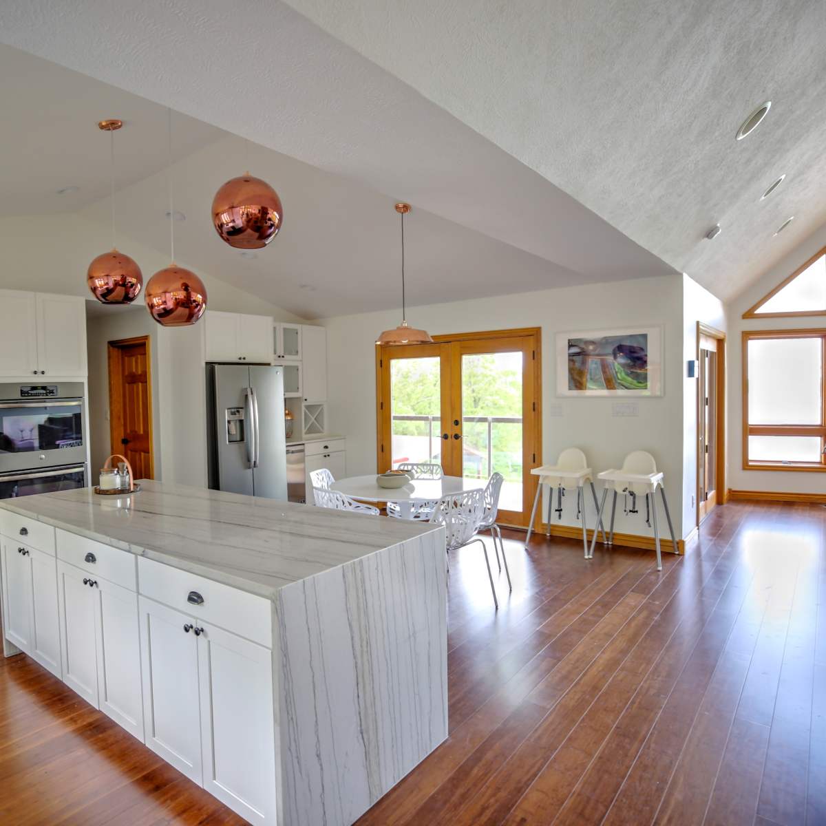 A modern kitchen with white cabinets, a marble island, and copper pendant lights, adjacent to a dining area with a table and chairs under large windows.