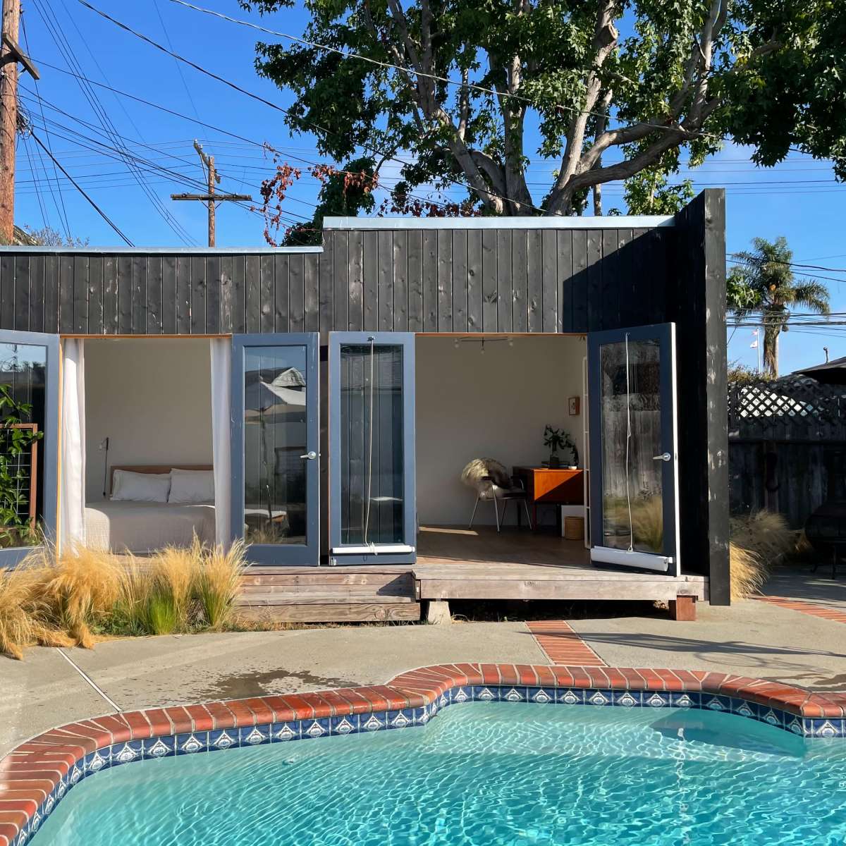A small modern structure with large glass doors sits beside a swimming pool, surrounded by greenery and utility poles against a clear blue sky.